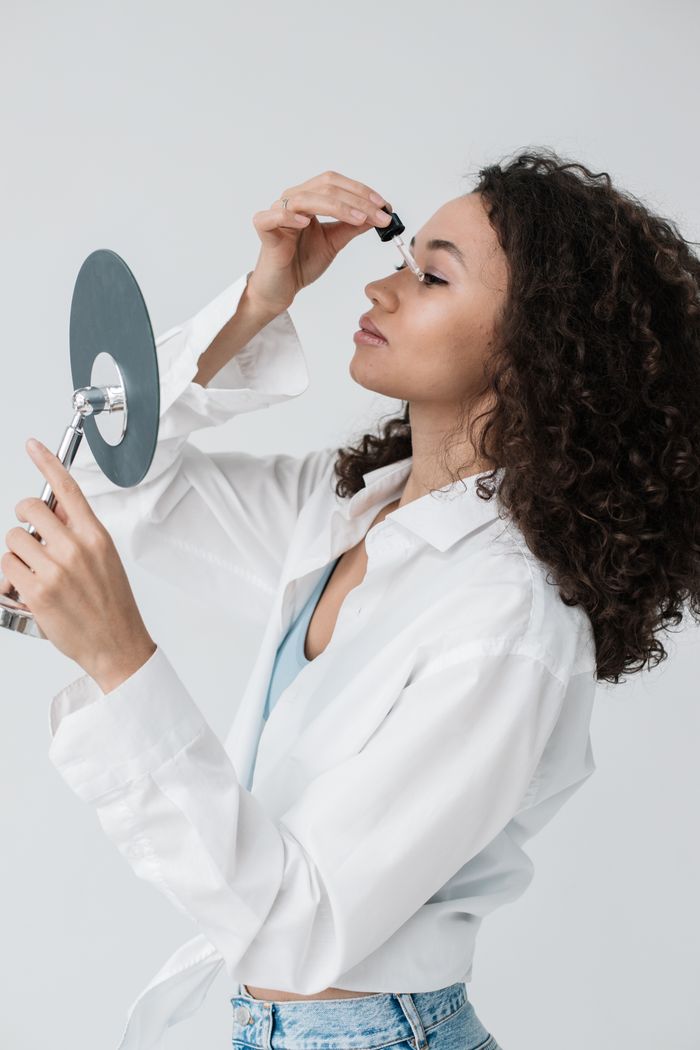A woman with curly hair applies skincare serum using a dropper in front of a mirror.